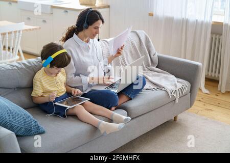 Freiberufliche Mutter sitzt auf dem Sofa im Home Office während Lockdown Arbeit auf Laptop, Kind spielt auf dem Tablet Stockfoto