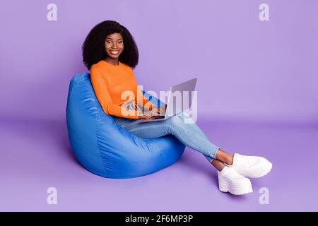 In voller Größe Profilfoto des optimistischen schönen Brünette Dame sitzen Typ Laptop tragen orange Top Jeans Sneaker isoliert auf Flieder Hintergrund Stockfoto