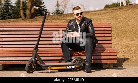 An einem Frühlingstag sitzt ein junger Mann auf einer Bank. Elektroroller neben einer Bank Stockfoto