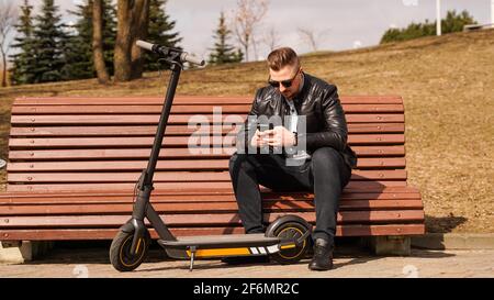 An einem Frühlingstag sitzt ein junger Mann auf einer Bank. Elektroroller neben einer Bank Stockfoto