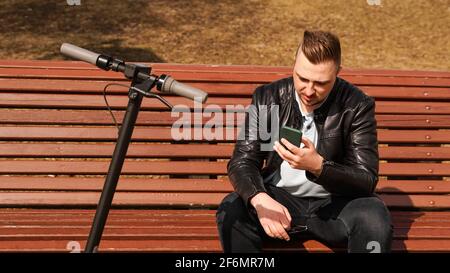 An einem Frühlingstag sitzt ein junger Mann auf einer Bank. Elektroroller neben einer Bank Stockfoto