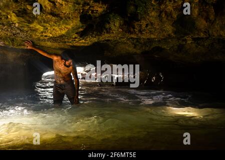 Attraktiver Mann mit Tattoos, der in den spektakulären Mermaid Caves mit Wellen auf vulkanischen Felsen steht, im Nanakuli Beach Park, Oahu, Hawaii, USA Stockfoto