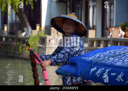 Eine Dame nimmt Touristen mit einem Kanalboot durch die Wasserstraßen von Zhouzhuang. In einheimischer Kleidung mit traditionellem Bambushut. Stockfoto