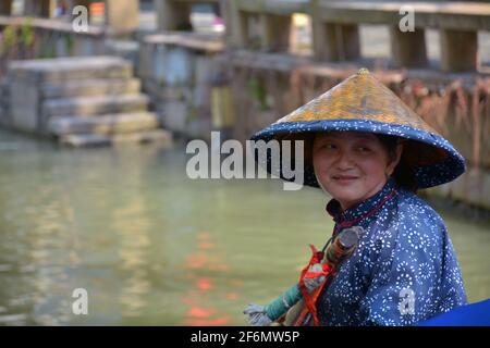 Eine Dame nimmt Touristen mit einem Kanalboot durch die Wasserstraßen von Zhouzhuang. In einheimischer Kleidung mit traditionellem Bambushut. Stockfoto