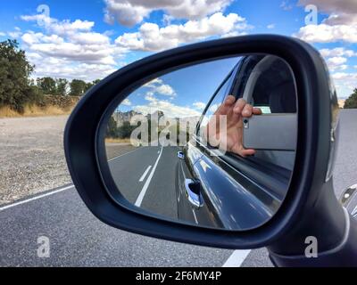 Spiegelung im Rückspiegel eines Autos eines Hand mit einem Handy, das ein Bild des nimmt Reflexion zeigt eine Straße mit einer Berglandschaft in Stockfoto