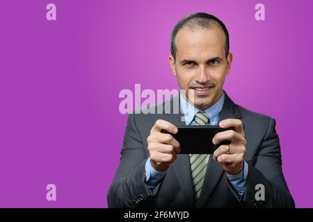 Reifer Mann mit Anzug und Krawatte, lächelnd und Smartphone waagrecht haltend. Stockfoto