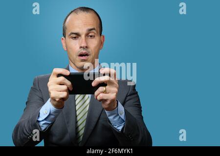 Reifer Mann mit Anzug und Krawatte, spielt auf seinem Smartphone und mit überraschendem Gesicht. Blauer Hintergrund. Stockfoto