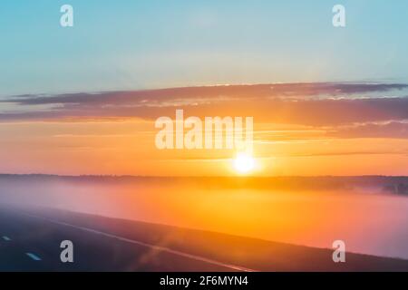 Morgengrauen mit Nebel, neblige Straße. Schöne Landschaft am frühen Morgen mit leuchtend orange aufgehenden Sonne und Wolken. Stockfoto