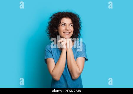 Nachdenkliche kaukasische lockige haarige Frau in einem blauen Kleid ist Träumen von etwas an einer Studiowand, das wegschaut Stockfoto