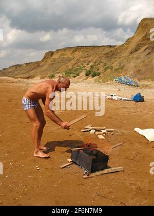 Ein gebräunter Mann verbrennt Holzbarren am Strand, einen Grill, einen Sonnenschirm, Hügel und Wolken am Himmel Stockfoto