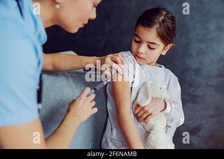 Tapferes kleines Mädchen, das zu Hause auf dem Sofa sitzt und Impfungen bekommt. Stockfoto