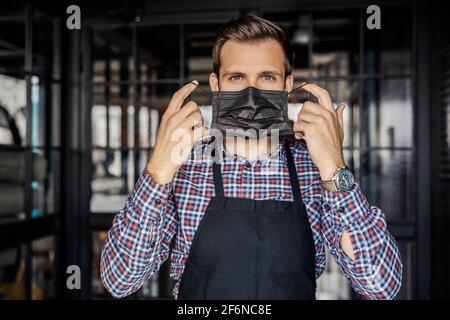 Ausziehen oder Aufsetzen einer Gesichtsmaske. Ein gutaussehender Kellner mit schönen Augen trägt eine Uhr am Handgelenk und steht am Eingang des Restaurants Stockfoto