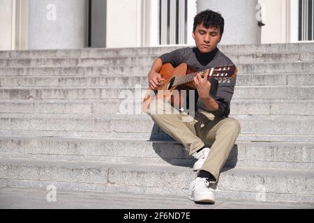 Lateinischer junger Mann, der Gitarre spielt und auf einer Treppe sitzt. Gitarre spielen im Freien. Stockfoto