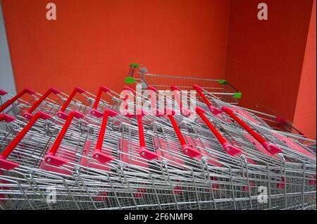 Reihen leerer metallischer Supermarktkarren im Freien auf der Straße in der Nähe des Ladens auf einem Hintergrund einer leuchtend orangefarbenen Wand. Stockfoto