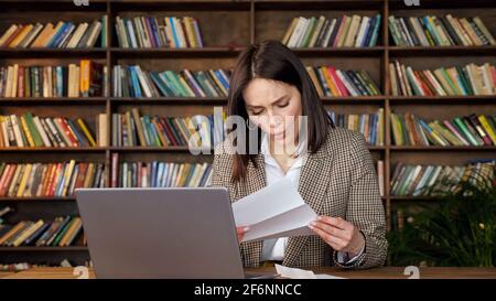 Junge Frau mit kurzen dunklen Haaren öffnet Bankbrief und Berührt die Wange mit einem Schock, der am Laptop gegen hölzerne Bücherregale sitzt Zu Hause Stockfoto