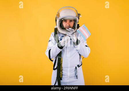 Schwul-transsexueller männlicher Kosmonaut mit ernsthafter Geste im Raumanzug und Helm, mit Transgender-Flagge, auf gelbem Hintergrund. Stockfoto