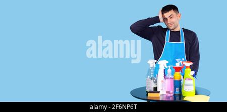 Banner, Langformat. Der junge Mann ist der Hausarbeit müde. Ein Mann hält seine Hand auf dem Kopf vor sich einen Holztisch mit unterschiedlicher Reinigung Stockfoto