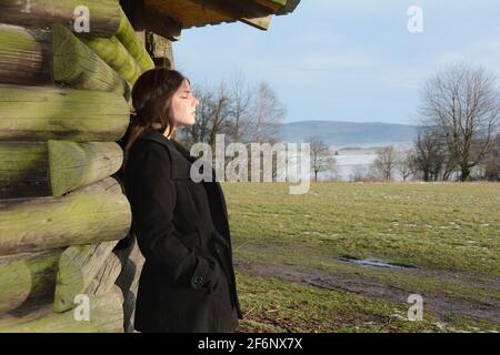 Junge Frau von der Seite, mit schwarzer Jacke, lehnt an eine Holzhütte, mit einem ernsten Ausdruck auf ihrem Gesicht und geschlossenen Augen, genießen die Sonne, ich Stockfoto