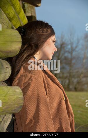 Junge Frau von der Seite, gelehnt an eine Holzhütte, mit einem ernsten Gesichtsausdruck und geschlossenen Augen, genießt die Sonne, in grüner Natur Stockfoto