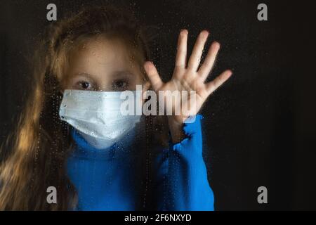 Baby Mädchen in medizinische Maske hinter nassem Glas.Tropfen auf Glas. Dunkler Hintergrund Stockfoto