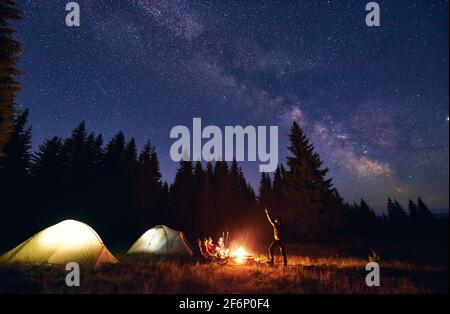 Mann zeigt seinen Freunden Milchstraße über Zeltstadt. Jungs sitzen am Feuer auf dem Hintergrund des Fichtenwaldes und genießen den Abend Sternenhimmel. Erholung im Freien. Nachtcamping in den Bergen Stockfoto