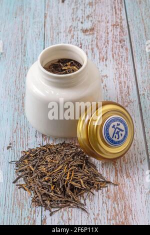 Teeblätter bereit für die Infusion und eine weiße Kanne, die Tee im Inneren enthält. Der Deckel, der auf dem Topf ruht, hat das Wort Tee auf Chinesisch geschrieben. Stockfoto