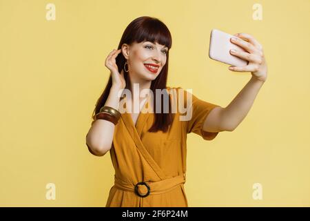 Aufgeregt in guter Laune, junge Frau, die trendigen gelben Anzug trägt, isoliert auf hellgelbem Hintergrund steht und Selfie auf dem Handy macht. Menschen, Technologien Konzept. Stockfoto