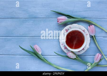 Dampfende heiße Tasse Tee und rosafarbene Frühlingsblumen über einem rustikalen blauen Holztisch, aufgenommen von oben. Stockfoto