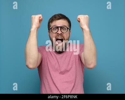 Ein emotionaler, glücklicher bärtiger Mann mit Brille und einem pinken T-Shirt macht eine Geste zum Sieger, schreit fröhlich und macht große Augen. Stockfoto