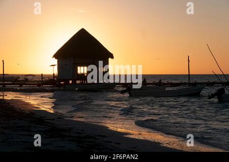 Eine kleine Fischerhütte auf einem hölzernen Pier über dem Meer bei Sonnenuntergang auf Holbox Island in Mexiko. Im Hintergrund sind der Himmel und Fischerboote. Reisetourismus Stockfoto