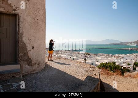 Ein junger Mann fotografiert von einem Hügel aus einen Überblick über das Dorf auf der Insel Mykonos in Griechenland. Im Hintergrund die Ägäis und die Küste Stockfoto