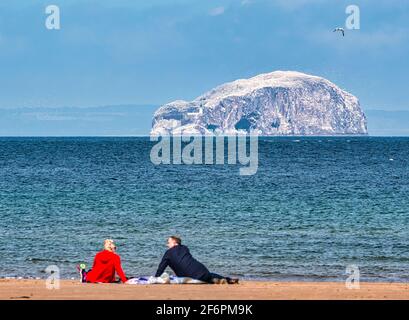 Ein Paar sitzt auf einer Picknickdecke am Strand von Belhaven Bay mit Blick auf die Bass Rock-Gannet-Kolonie an einem sonnigen, luftigen Tag, East Lothian, Schottland, Großbritannien Stockfoto
