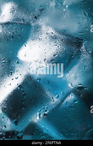 Eiswürfel in einem vernebelten Glas mit Tropfen Eiswasser Nahaufnahme Makro. Erfrischendes und kühlendes Getränk bei heißem Wetter. Blauer Hintergrund. Vollbild Stockfoto