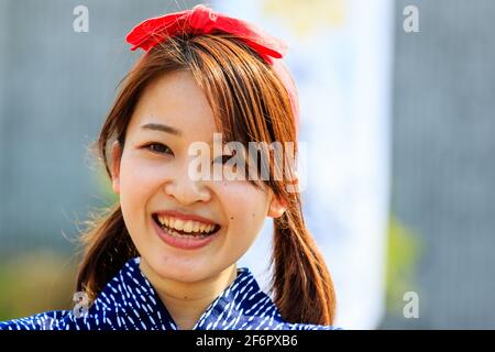 Porträt einer sehr hübschen japanischen jungen Frau, einer Yosakoi-Tänzerin, die im Freien steht und den Betrachter direkt anschaut und lächelt. Rote Schleife in ihrem braunen Haar. Stockfoto