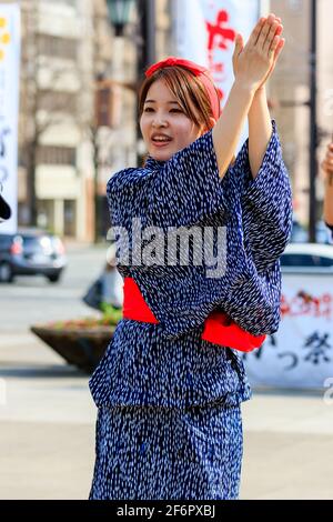 Japanische junge Frau, Yosakoi-Tänzerin, steht draußen auf der Straße mit erhobenen Händen klatschend. Trägt eine blaue Yukata mit einer roten Schärpe. Stockfoto