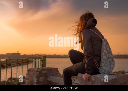 Junge Frau mit braunem Haar, die einen Rucksack trägt und die anschaut Horizont bei Sonnenuntergang am Meer Stockfoto