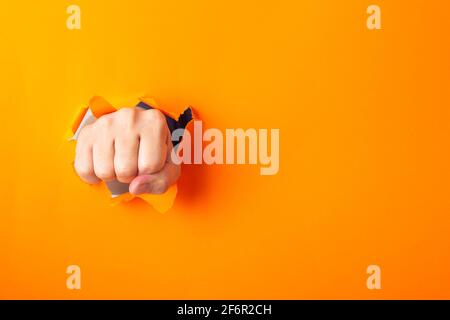 Die Faust brach oranges Papier und riss ein Loch. Durchstanzen Sie das Papier. Stockfoto