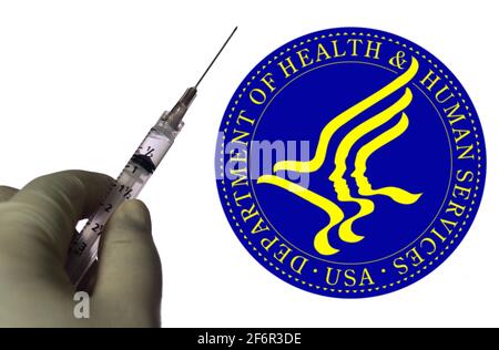 In dieser Abbildung hält eine Hand eine medizinische Spritze mit einem Unternehmenslogo des US-Gesundheitsministeriums (HHS), das auf einem Bildschirm im Hintergrund angezeigt wird. Stockfoto