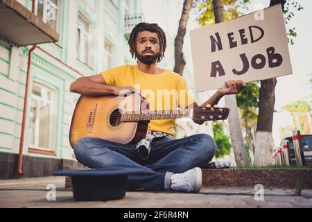 Foto-Portrait eines traurigen mürrischen Mannes, der die Gitarre im Sitzen hält Straße mit Karton auf der Suche nach Job Kopfbedeckung für Geld Stockfoto