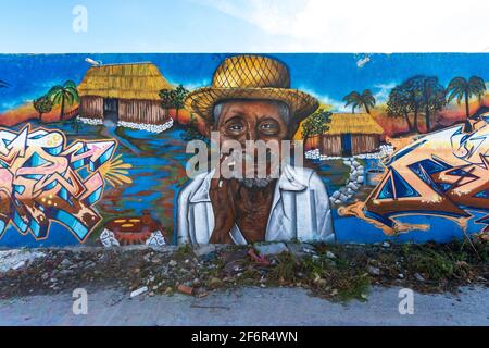 Street Art in einer kleinen Stadt an einer langen Wand mit einem alten Mann, der einen Strohhut, eine rustikale Hütte und Worte trägt, mit einem blauen Himmel über der Wand Stockfoto