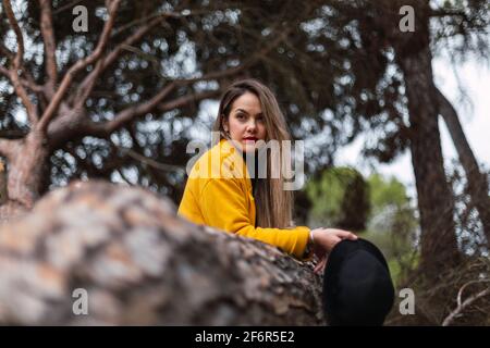 Schöne Frau, die sich mit einem gelben Mantel an einen Baum lehnt Und einen schwarzen Hut Stockfoto