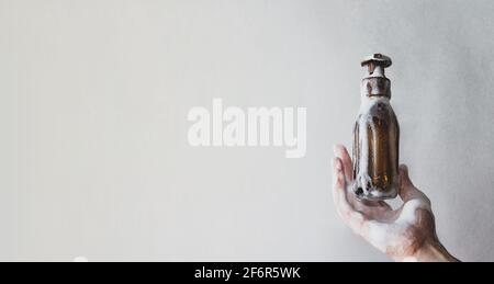 Frau hält eine Flasche Duschgel in Schaumstoff. Wiederverwendbare Glasflasche mit Spender für Shampoo, Seife oder Duschgel auf grauem Hintergrund. Stockfoto