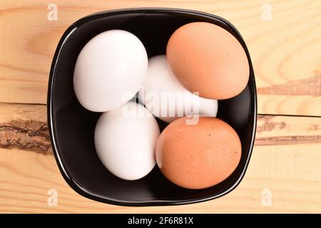 Mehrere beige und weiße frische Bio-Hühnereier in einer schwarzen Keramikplatte auf einem natürlichen Holzhintergrund. Stockfoto