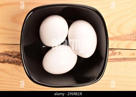 Drei frische, leckere, weiße Hühnereier aus biologischem Anbau in einer schwarzen Keramikplatte auf einer Arbeitsplatte aus natürlichem Holz. Stockfoto