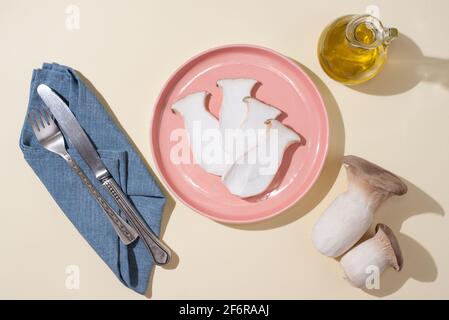 Rosafarbener Teller mit frischen Austernpilzen, Rosmarinzweigen, Messer, Gabel auf gelbem Hintergrund bei Sonnenlicht, Nahaufnahme. Stockfoto
