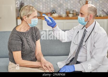 Ein Arzt misst die Temperatur einer älteren Frau in ihrem Zuhause mit einer Schutzmaske. Coronovirus-Infektionsdiagnosekonzept Stockfoto