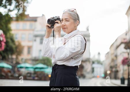 Kaukasische Rentnerin mit Retro-Kamera für die Aufnahme von schönen Architektur in der Altstadt. Ziemlich reife Frau, die Freizeit zum Reisen verbringt. Stockfoto