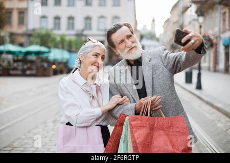 Fröhliches Seniorenpaar, das Selfie auf einem modernen Smartphone nimmt, während es mit Einkaufstaschen in den Händen auf der Straße steht. Konzept von Familie, Technologie und Kauf. Stockfoto