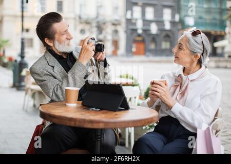 Reifer bärtiger Mann, der mit einer Retro-Kamera Fotos von seiner hübschen Frau machte. Glückliches Paar in stilvoller Kleidung, das sich auf der Cafeterrasse mit Tablet und Kaffee entspannt. Stockfoto
