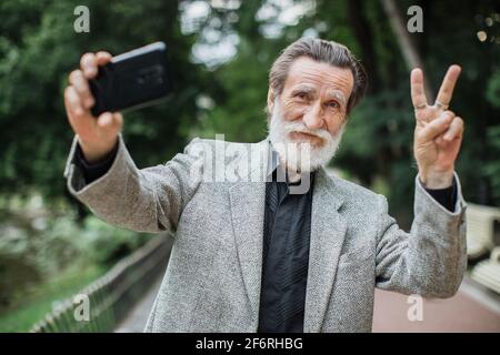 Stilvoller älterer Mann, der im grünen Park steht und Selfie auf einem modernen Smartphone nimmt. Glücklicher bärtiger Rentner, der vor der Kamera gestikulierte und lächelte. Stockfoto